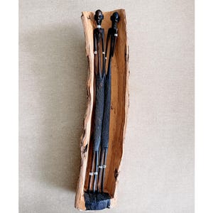 May include: A wooden sculpture featuring two slender, dark-toned figures with detailed carved features. They stand within a natural wood trough, showcasing a blend of textures and tones. The figures have silver-colored accents around their necks and arms.