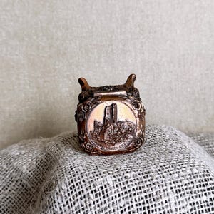 May include: A small, metallic, copper-toned trinket box with a hinged lid. The front of the lid features a raised relief design of a castle ruin surrounded by floral motifs, with the text RUINE DRACHENFELS embossed along the bottom edge.
