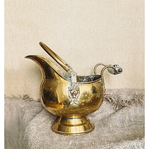 Pode incluir: Um balde de carvão em latão polido com um bico largo, uma alça arqueada resistente e uma base de pedestal. O corpo é decorado com um relevo de cabeça de leão e inclui uma pequena pega com um inserto decorativo de cerâmica.