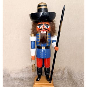 Könnte beinhalten: Eine hölzerne Nussknacker-Figur mit einem schwarzen Hut mit Goldrand, einer blauen Jacke mit goldenen Akzenten und einer roten Hose. Die Figur hat einen Bart und Haare aus braunem Kunstfell, hält einen schwarzen Speer und eine kleine goldene Laterne.