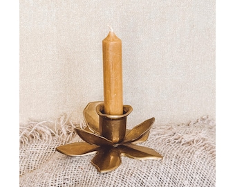 Puede incluir: Un portavelas de latón con forma de flor con seis pétalos que rodean una copa cilíndrica central. Una vela cónica única, lisa y de color marrón claro está insertada en el centro del soporte, manteniéndose erguida dentro de la estructura.