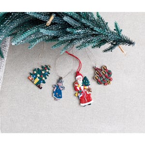 Puede incluir: Cuatro adornos navideños colgantes que incluyen un árbol verde con regalos, un ángel azul con estrellas, un Santa Claus cargando un árbol y un saco, y una corona circular con tres velas. Cada adorno cuelga de un hilo fino sobre un fondo neutro.