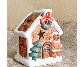 May include: A ceramic gingerbread house figurine featuring white icing trim, a red bow, a green wreath, a star-shaped cutout, a green tree, and a gingerbread person figure. The house has a brown textured finish and a red heart cutout on the side.