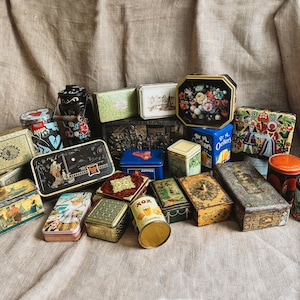 Pode incluir: Uma coleção de latas de metal vintage em várias formas e tamanhos. As latas apresentam desenhos florais, paisagísticos e geométricos, com cores como dourado, azul e vermelho. Algumas latas têm texto, tudo disposto sobre um fundo de tecido neutro.