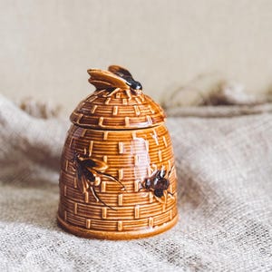 May include: A brown ceramic beehive-shaped jar with a lid, featuring a bee figurine on top. The jar has a woven texture and bee illustrations on the sides. This decorative item is suitable for storing honey or other small items.