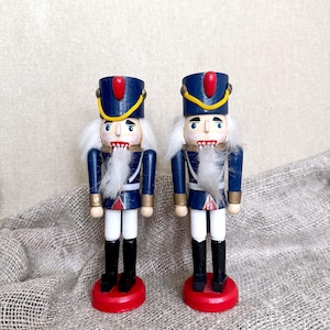 May include: Two wooden nutcrackers in a classic soldier design. They feature blue jackets, white hair, and black boots. Each stands on a red base, with gold and white accents. The nutcrackers are approximately 10 inches tall.