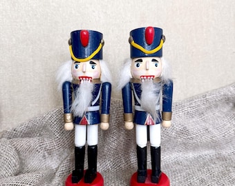 May include: Two wooden nutcrackers in a classic soldier design. They feature blue jackets, white hair, and black boots. Each stands on a red base, with gold and white accents. The nutcrackers are approximately 10 inches tall.