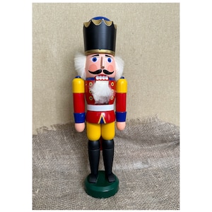 Peut inclure: Un casse-noisette en bois traditionnel avec un uniforme rouge, jaune et bleu. Le casse-noisette porte un chapeau noir avec une couronne dorée, une barbe blanche et une moustache. Le casse-noisette est debout sur une base verte.