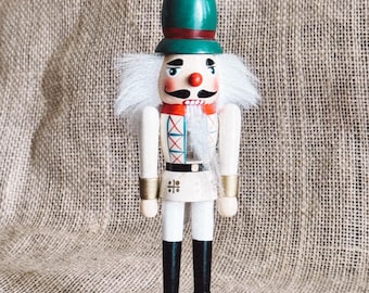 May include: A traditional wooden nutcracker with a white beard, green hat, and red and white accents. The nutcracker is standing on a red base.