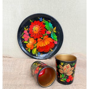 Könnte beinhalten: Ein schwarzer runder Teller und zwei passende Becher mit handgemalten Blumenmustern in leuchtendem Rot, Orange und Grün. Der Teller zeigt ein großes zentrales Blumenarrangement, während die Becher goldfarbene Innenseiten und florale Motive haben.