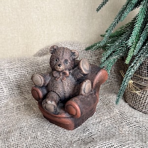 Op de afbeelding: Een bruin teddybeerbeeldje dat in een miniatuur bruine fauteuil zit. De beer heeft een strikje en is gemaakt van een textuurrijk materiaal. Een kleine decoratieve kerstboom staat op de achtergrond.