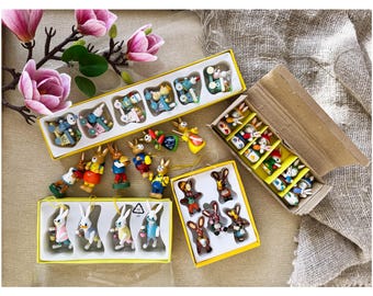 May include: A collection of small, hand-painted wooden rabbit figurines in various poses and outfits, some packaged in yellow or cardboard boxes with white plastic inserts, and others arranged loosely on a surface.