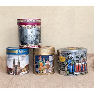 May include: Four cylindrical metal storage tins with decorative lids. One tin is red and silver with a festive scene, one is blue with a cathedral illustration, one is gold with a craftsman graphic, and one is silver with a medieval royal scene.