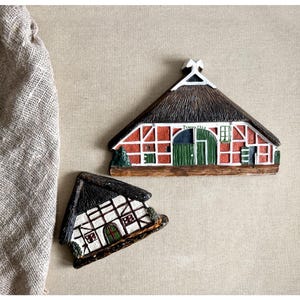 Può includere: Due calamite a forma di casa in miniatura. Una è rossa con tetto di paglia e porta verde, l'altra è bianca con tetto di paglia nero. Le calamite sono dettagliate con finestre e finiture decorative, evocando un tema rustico.