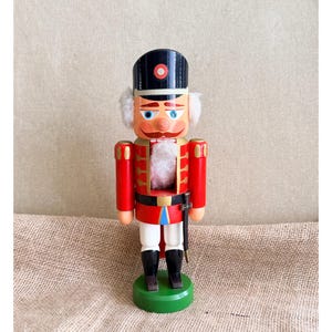 May include: A wooden nutcracker figurine wearing a black hat with a red and white circular emblem, a red jacket with gold trim, white trousers, and black boots. It features white faux fur hair and a beard, blue eyes, and a small sword at its side.