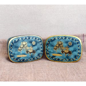 May include: Two oval-shaped metal boxes featuring a mottled turquoise blue finish with gold-coloured trim. The lids display a black graphic illustration of a person riding a donkey near palm trees, accented by a gold sun and a zigzag border pattern.