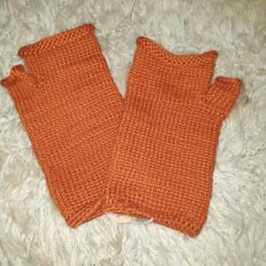 May include: A pair of orange fingerless gloves, knitted with a textured stitch. The gloves are made of a warm, soft material and are designed to cover the hand while leaving the fingers free. The gloves are displayed on a white, fluffy surface.