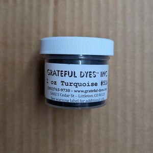 May include: A small, clear plastic jar with a white lid, filled with turquoise dye. The label reads "GRATEFUL DYES INC" and "1 oz Turquoise #511". The jar is on a light brown surface.