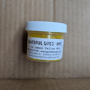 May include: A small, clear plastic jar with a white lid, filled with bright yellow powder. The label reads "GRATEFUL DYES&trade; INC." and "1 oz Lemon Yellow #38." Additional text includes a phone number and website address.