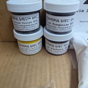 May include: Four small jars of dye are stacked. The jars have white lids and black labels with text. The labels indicate the colors are Ultra Violet, Turquoise, Lemon Yellow, and Fuchsia. The jars are labeled "Grateful Dyes Inc."
