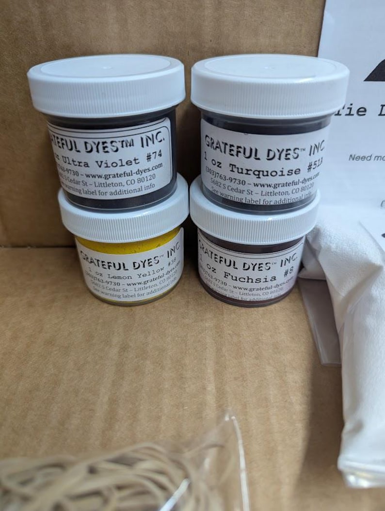 May include: Four small jars of dye are stacked. The jars have white lids and black labels with text. The labels indicate the colors are Ultra Violet, Turquoise, Lemon Yellow, and Fuchsia. The jars are labeled "Grateful Dyes Inc."