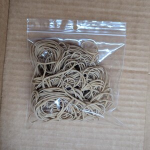 May include: A clear plastic bag filled with a large quantity of tan rubber bands. The rubber bands are tightly packed together, creating a tangled mass. The bag is sealed and sits on a brown cardboard surface.