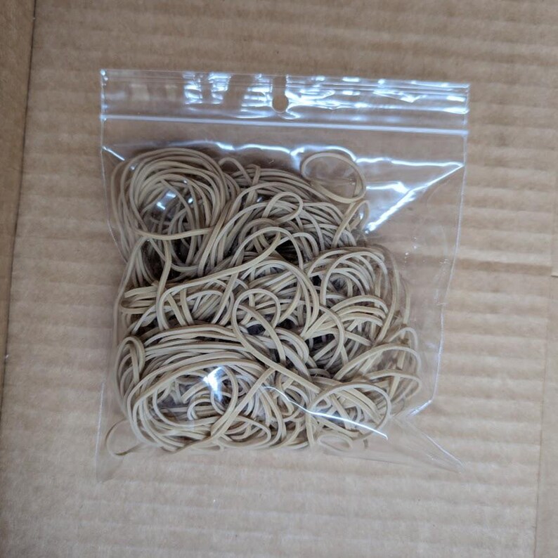 May include: A clear plastic bag filled with a large quantity of tan rubber bands. The rubber bands are tightly packed together, creating a tangled mass. The bag is sealed and sits on a brown cardboard surface.