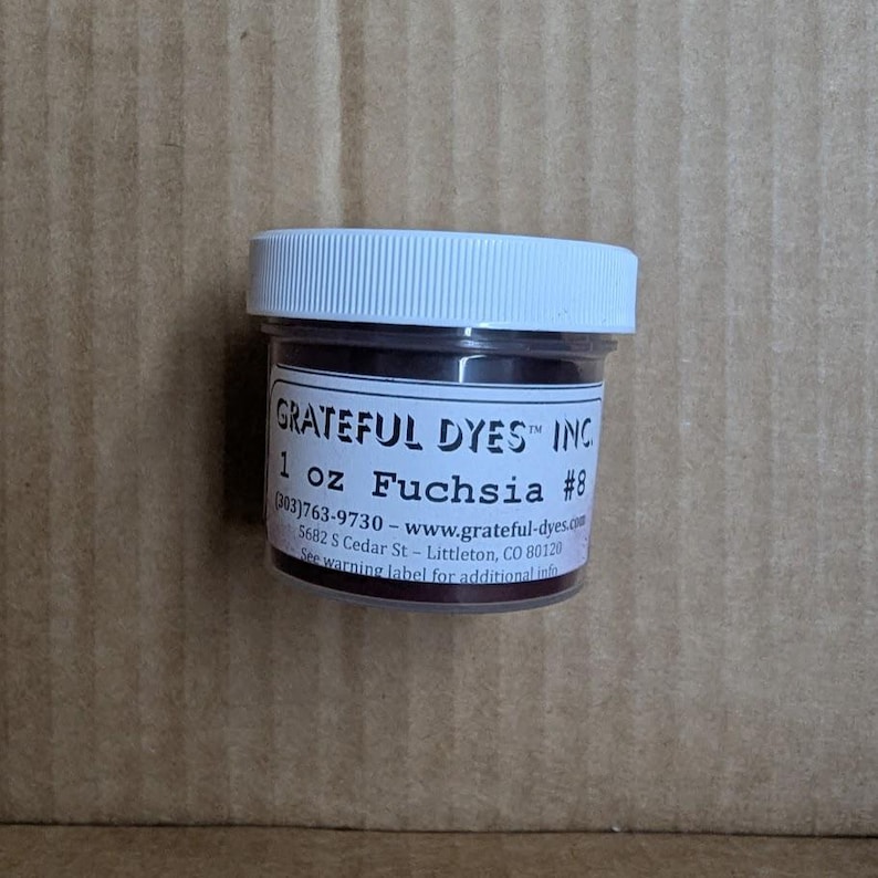 May include: A small, clear jar with a white lid containing fuchsia-colored dye. The label reads "GRATEFUL DYES INC" and "1 oz Fuchsia #8". The jar is on a brown cardboard surface.