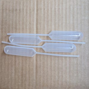 May include: Four clear plastic pipettes with bulb-shaped reservoirs and thin, elongated tips are arranged on a light brown, textured surface. The pipettes are designed for precise liquid transfer in a laboratory or scientific setting.