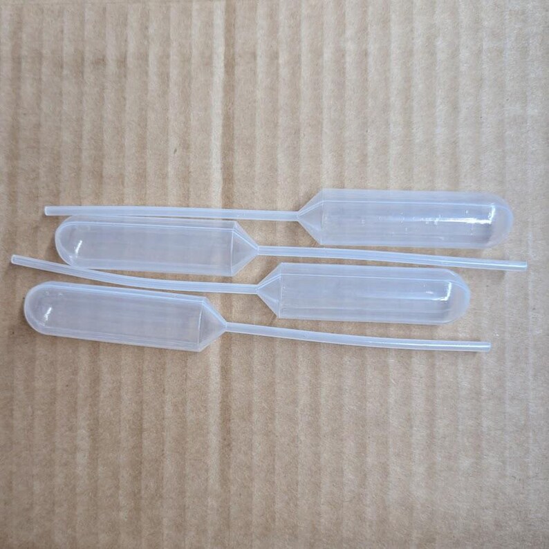 May include: Four clear plastic pipettes with bulb-shaped reservoirs and thin, elongated tips are arranged on a light brown, textured surface. The pipettes are designed for precise liquid transfer in a laboratory or scientific setting.