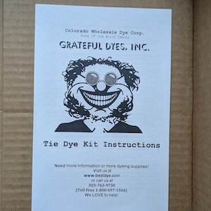 May include: A white instruction sheet for a tie-dye kit. The sheet features the text "Grateful Dyes, Inc." and a cartoon face with sunglasses and a large grin. Additional text includes website and phone number.