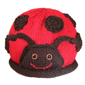 Può includere: Cappello di coccinella rosso a maglia con macchie nere e un viso sorridente. Il cappello ha un bordo a maglia nero e occhi marroni.