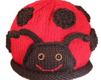 May include: Red knitted ladybug hat with black spots and a smiling face. The hat has a black knitted brim and brown eyes.