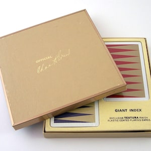 May include: A gold box with a gold lid. The lid has the text "OFFICIAL" and a signature that says "Charles Atlas". The box contains a deck of playing cards with a red and white back design. The box has the text "GIANT INDEX EXCLUSIVE TEXTURA FINISH PLASTIC COATED PLAYING CARDS".
