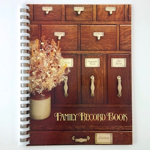May include: A brown spiral bound book with a vintage aesthetic. The cover features a photo of wooden drawers with gold hardware and a bouquet of dried flowers in a cream-colored vase. The text "FAMILY RECORD BOOK" is printed in gold lettering on the cover.