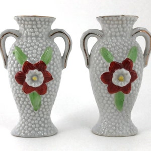 May include: Pair of small white ceramic vases with gold handles and red flower designs. The vases have a textured surface.