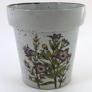May include: A ceramic flower pot with a light gray speckled finish. The pot features a floral design with purple and yellow flowers and green leaves. The rim of the pot is a darker gray.