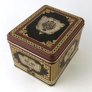May include: A rectangular metal storage tin with a hinged lid. The tin features a dark brown background with ornate gold and black decorative elements. The lid and sides have a repeating pattern and a central emblem.