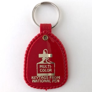 May include: A red plastic keychain with a silver key ring. The keychain has a teardrop shape with a gold emblem that reads "MULTI-COLOR KEYTAGS FROM NATIONAL PEN". The edges are textured.