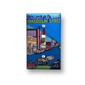 May include: A light switch cover featuring a colorful illustration of a street scene with the text "Grateful Dead Shakedown Street".