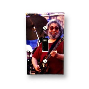 May include: A light switch cover featuring a color photograph of a man with a beard and sunglasses playing a guitar. The man is wearing a red shirt and is standing in front of a drum set.