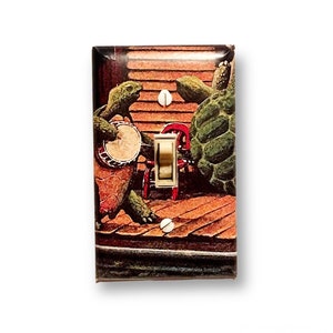 May include: A light switch cover featuring a vintage illustration of two turtles playing music. The turtles are green and brown, and the background is a wooden porch with a red chair.