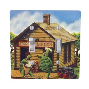 May include: A decorative light switch cover featuring a whimsical illustration. Two green turtles are dancing on a wooden porch in front of a building labeled "Terra Station." The background includes a blue sky and trees.