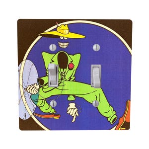 Peut inclure: Un cache-interrupteur d&eacute;coratif avec un personnage de dessin anim&eacute; en costume vert et chapeau jaune sur fond bleu. Le motif comprend une fleur rouge et une horloge. Le couvercle comporte deux interrupteurs argent&eacute;s.