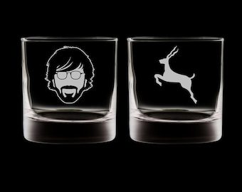 May include: Two clear, square-shaped whiskey glasses. One glass features a white etched image of a man's face with glasses, a beard, and mustache. The other glass has a white etched image of a leaping deer.