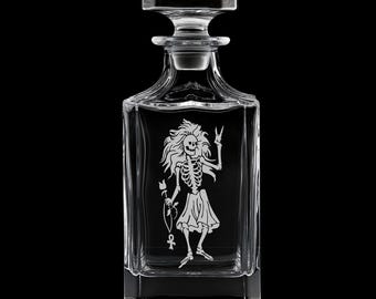 May include: A clear glass decanter with a square base and a stopper. The decanter features an etched design of a skeleton figure with long hair, holding a flower and giving a peace sign. The background is black.