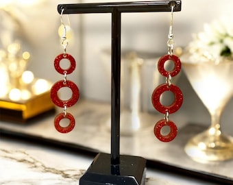 May include: A pair of dangling earrings with three graduated red glittery circles suspended from silver-colored hooks. The earrings are displayed on a black stand against a blurred background.
