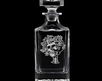 May include: A clear glass decanter with a square base and a stopper. The decanter features an etched design of a skull surrounded by roses. The design is centered on the front of the decanter. The background is black.