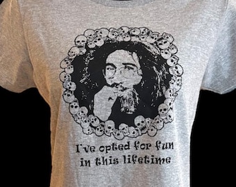 May include: A light gray t-shirt featuring a black and white portrait surrounded by skulls. The text "I've opted for fun in this lifetime" is printed below the portrait. The shirt is displayed on a black mannequin.