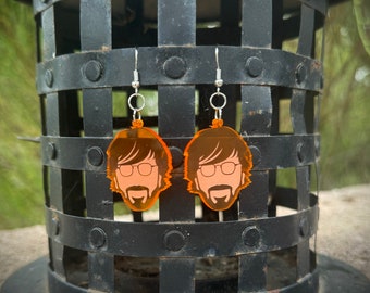 May include: A pair of orange acrylic earrings featuring a cartoon face with glasses and a beard. The earrings are hanging from silver hooks.